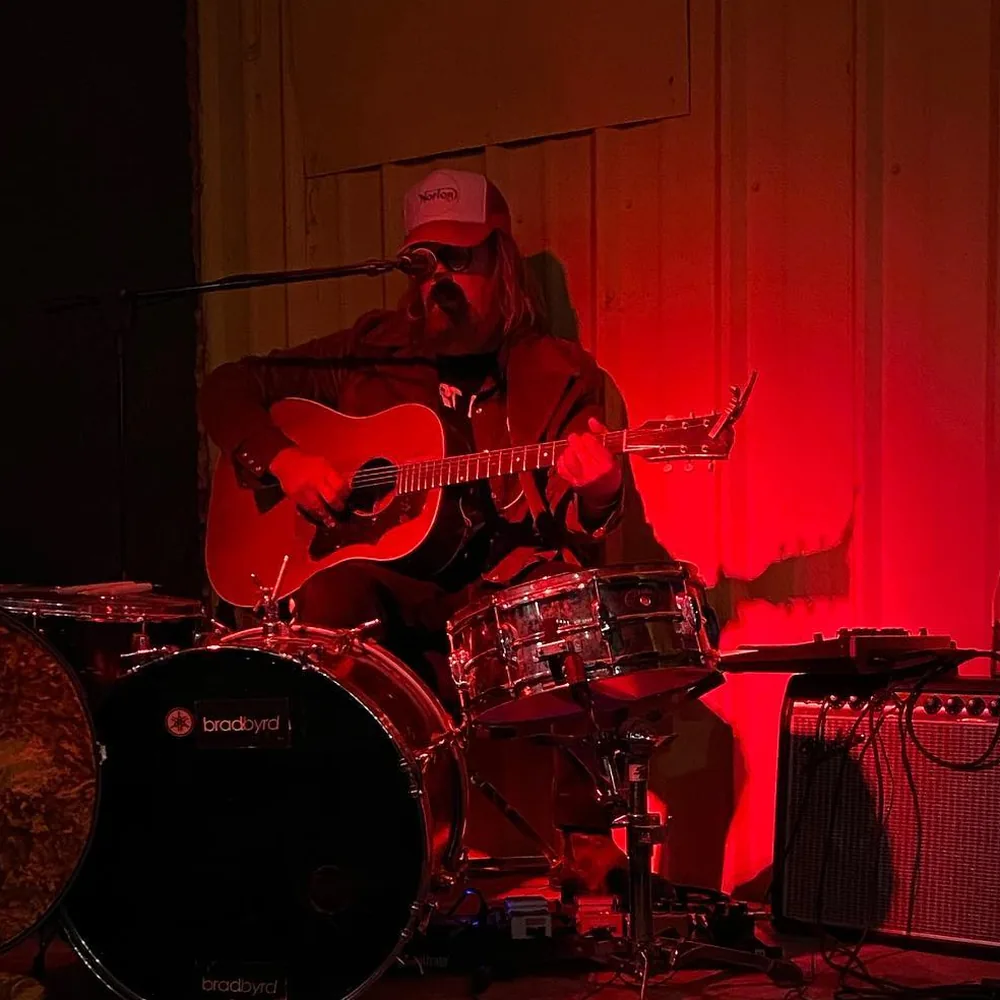 Brad Byrd playing acoustic guitar alone behind a kick drum and snare under red light at Giant Rock Meeting Room